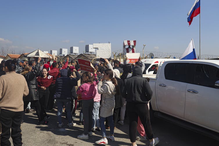 Several people, including women and children, stand next to parked vehicles.