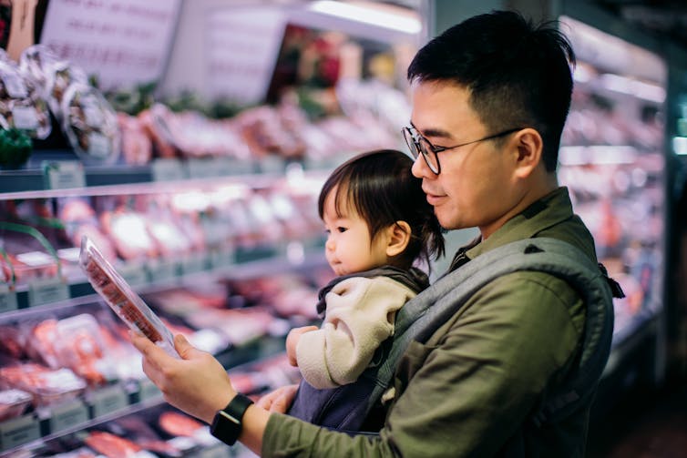 Man with baby in carrier looks at meat in supermarket.