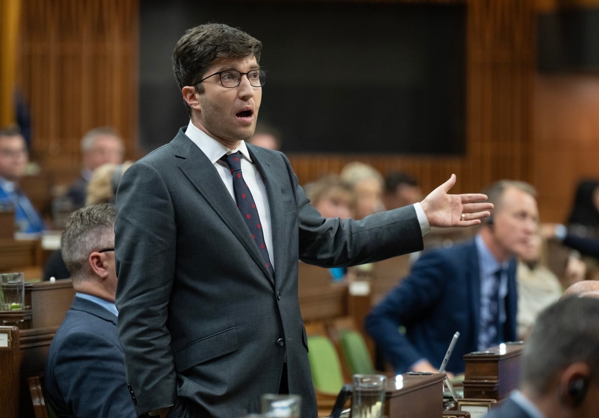 A man in a grey suit gestures with his left hand as he stands and speaks in the House of Commons.