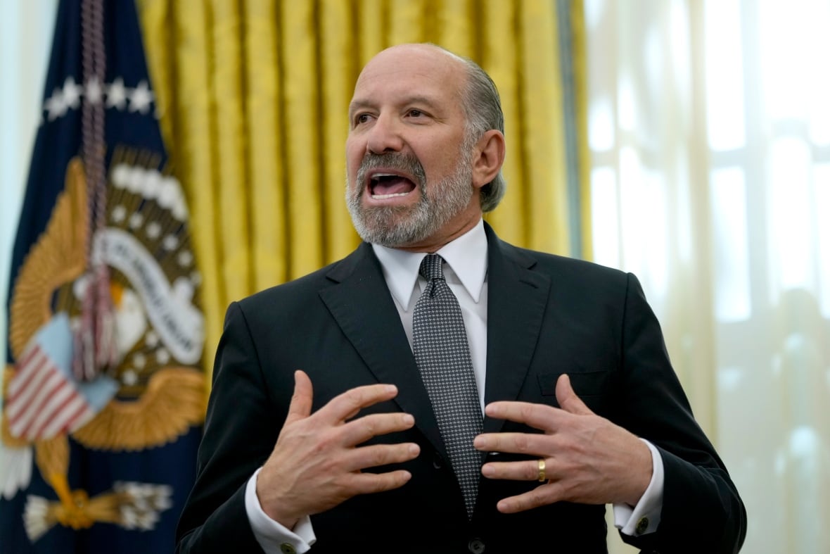 A man in a dark suit speaks while gesturing with his hands. 