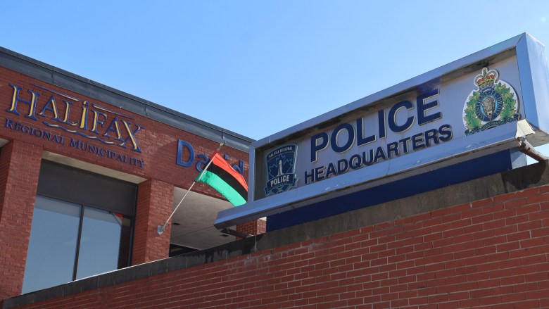 A steel sign that says Police Headquarters with a blue and white logo with a lighthouse and Halifax Regional Police to the eft and another logo with a crown to the right stands atop a red brick wall. A red, black, and green striped flag hangs over the front door while another sign that says Halifax Regional Municipality is mounted on the front of the building.
