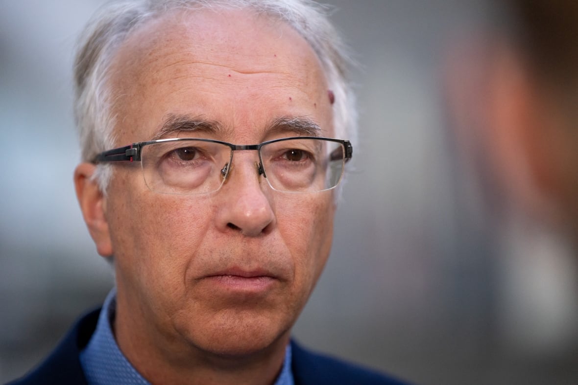 A man with white hair and glasses wearing a suit and blue collared shirt is pictured in a close-up.