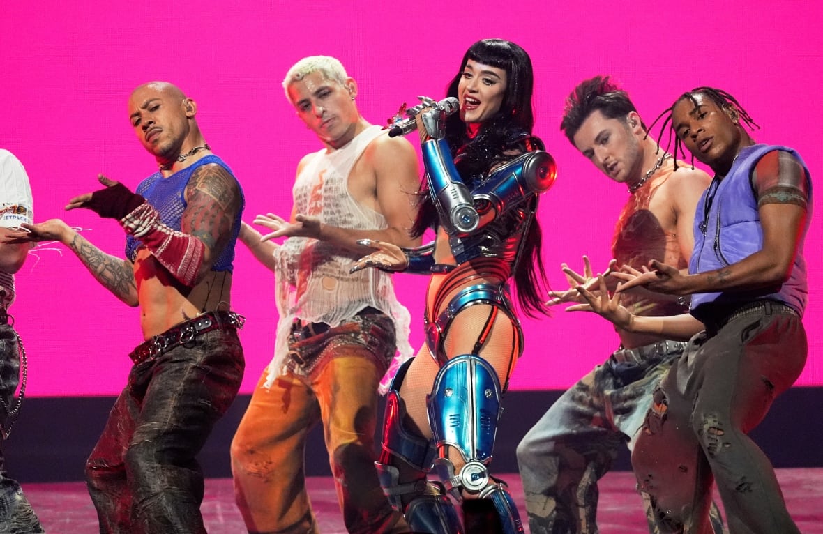 A woman wearing a metallic blue outfit holds a microphone and sings on stage as men dance on either side of her. 