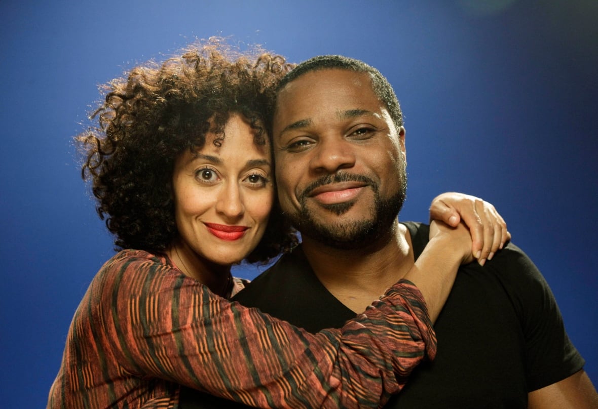 A woman with a striped blouse and curly hair wraps he arms around the shoulders of a man wearing a black t-shirt.