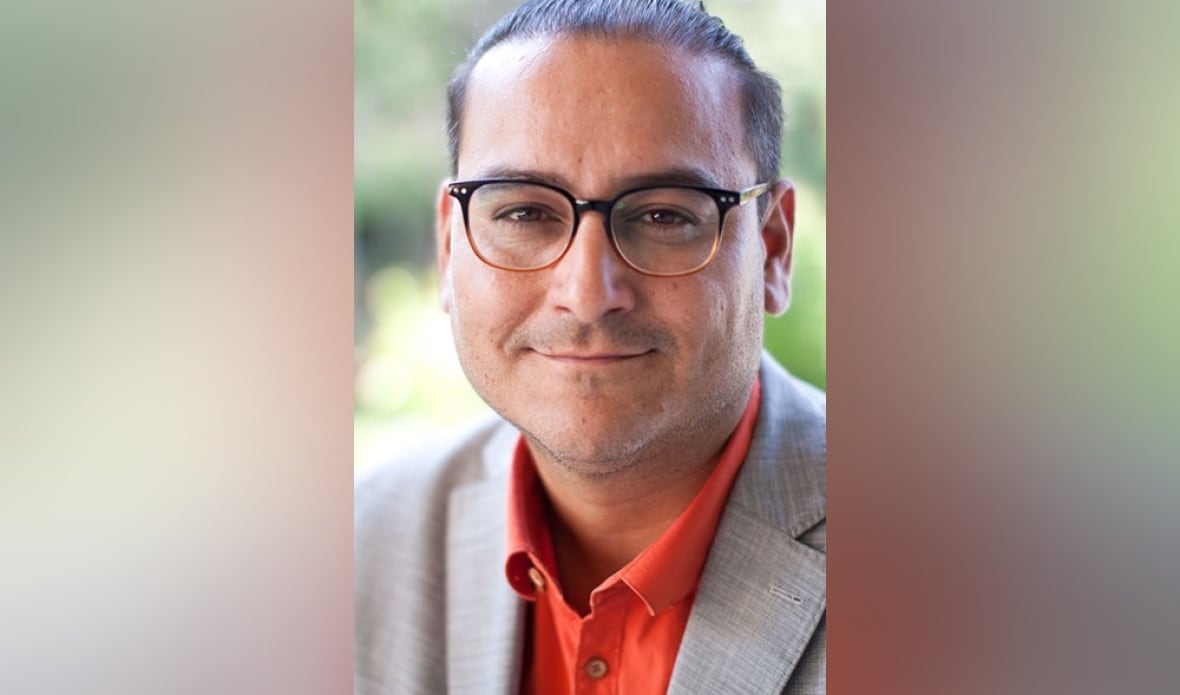 A man wearinghis hair in a ponytail, with a suit and glasses, smiles to the camera.