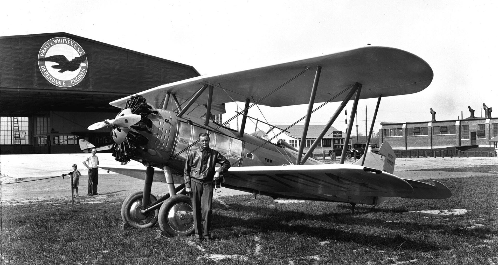 black and white photo of a man in front of a biplane and a hangar is in the background