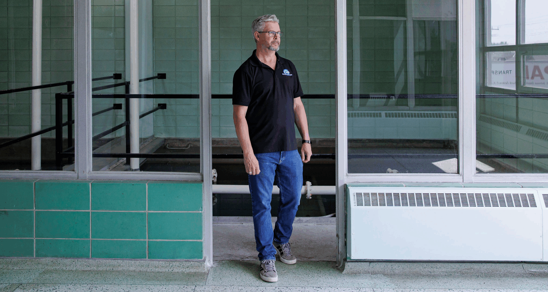 A man wearing a black polo shirt, jeans and sneakers walks through an interior doorway. He has short grey hair and a trimmed beard. He is wearing glasses. There is green tile on the wall behind him and a staircase leading down. Either side of the doorway is panelled glass.