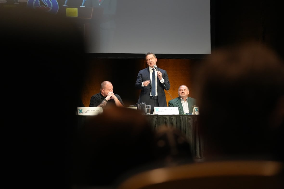 A man speaking beside two people seated at a table. The silhouettes of people from a crowd can be seen as well. 