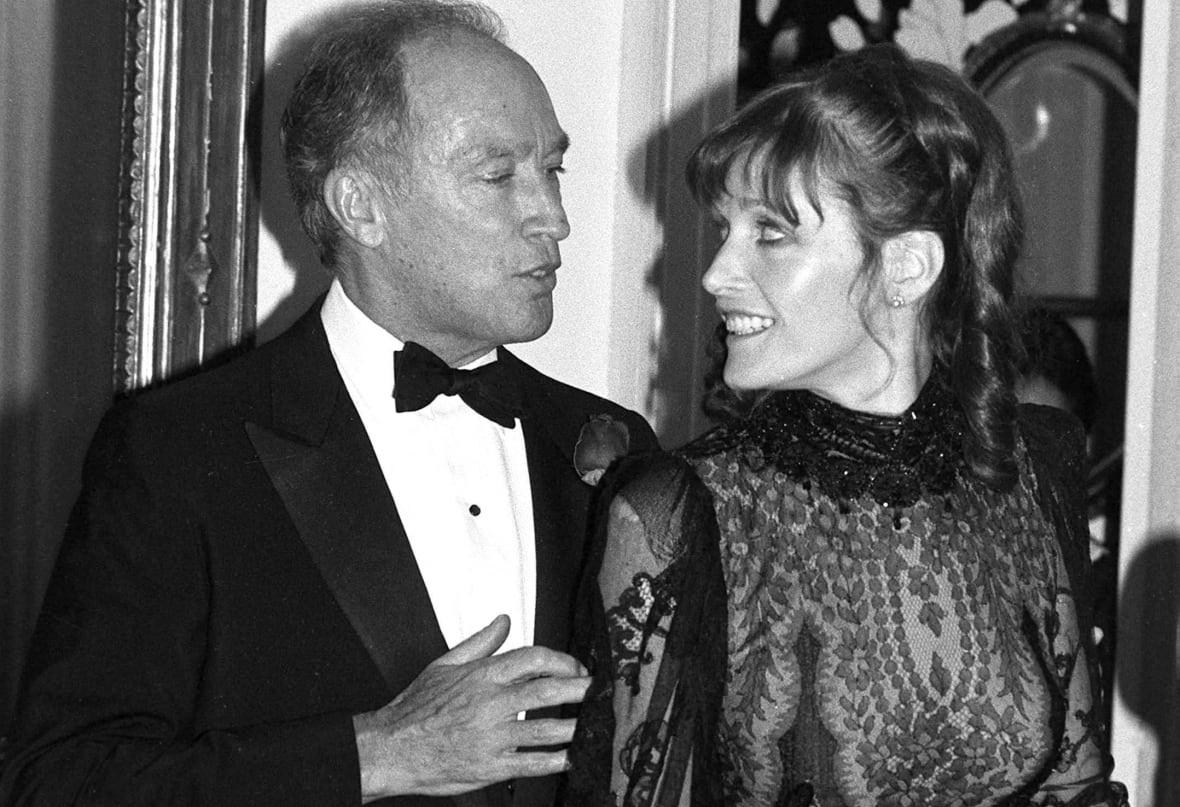 A black and white photograph of a man wearing a black tuxedo, black bowtie and white shirt walking with his arm behind a woman, on his right, who is wearing a black gown. 