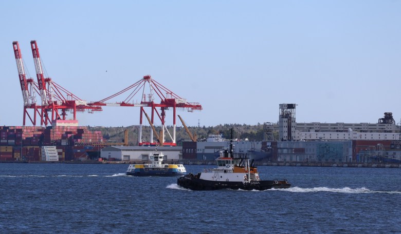 A ferry and a tugboat travelling in opposite directions in a working harbour in front of a port with tall red and white cranes and a grain elevator to the right. There are stacks of colourful shipping containers at the left.