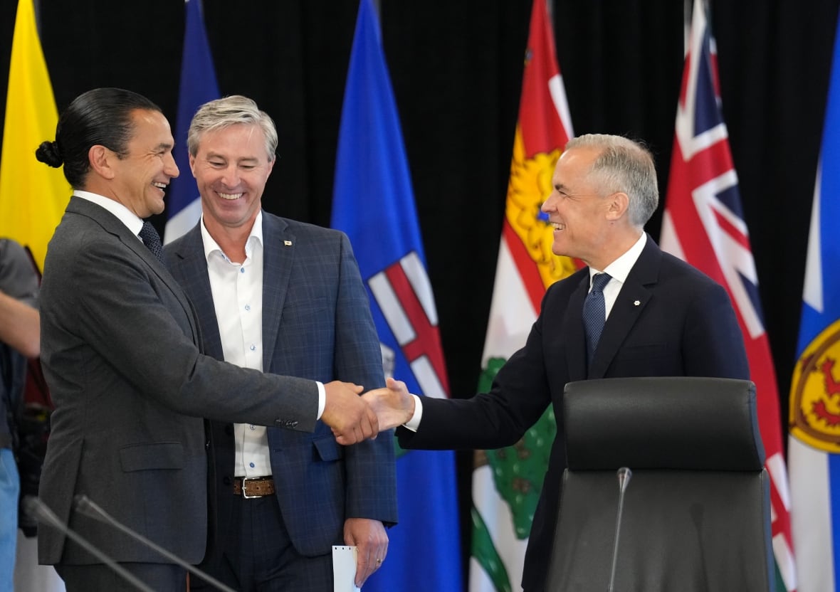 Three men chat in front of a table and flags.