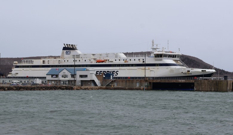 A car and passenger ferry painted in white with "Bay Ferries" written on the side is docked at a concrete wharf. There is a small terminal in front of the wharf and cars parked in the area.