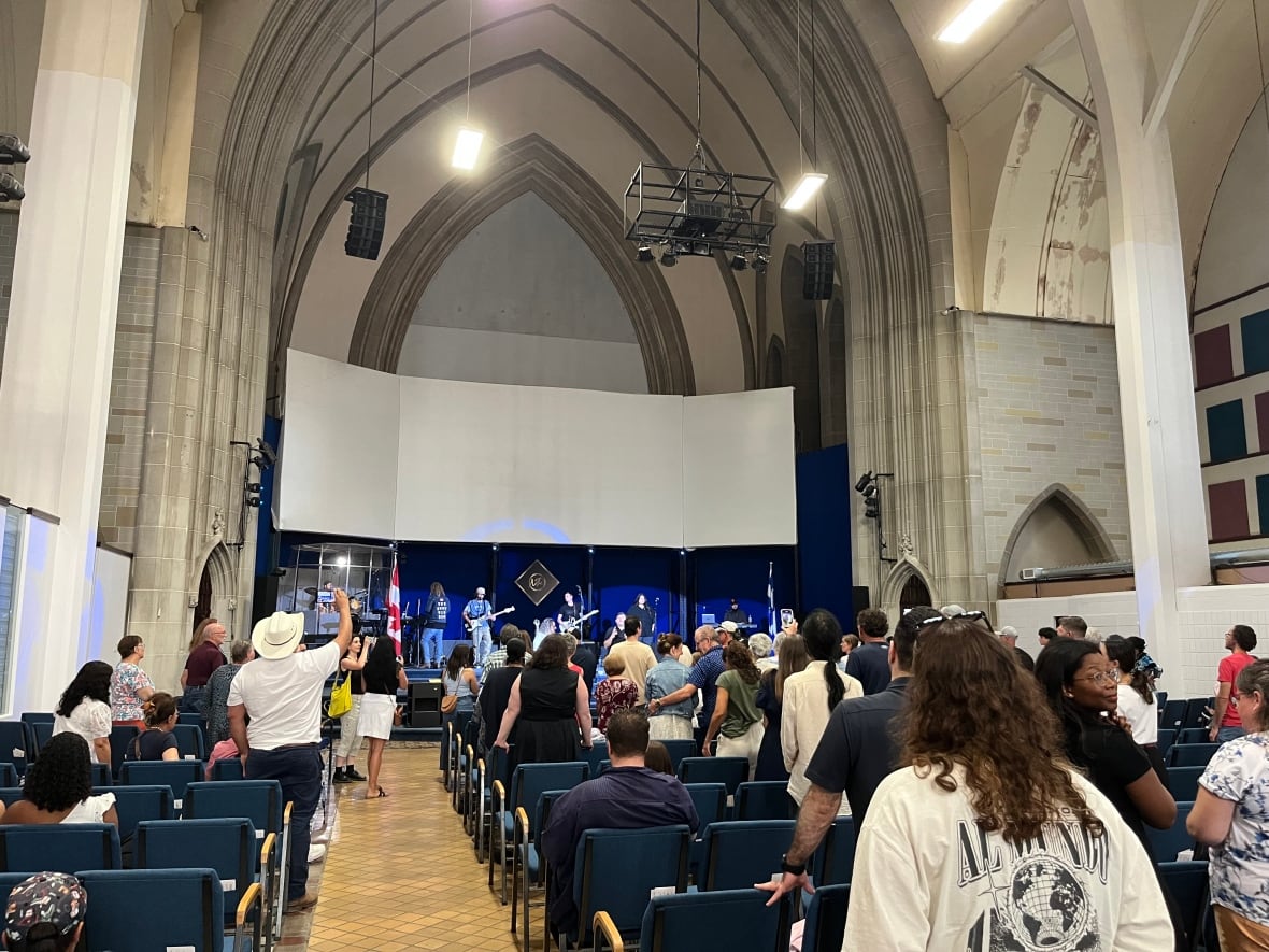 Musicians appear on a stage inside a church with worshippers sitting or standing as they look on.