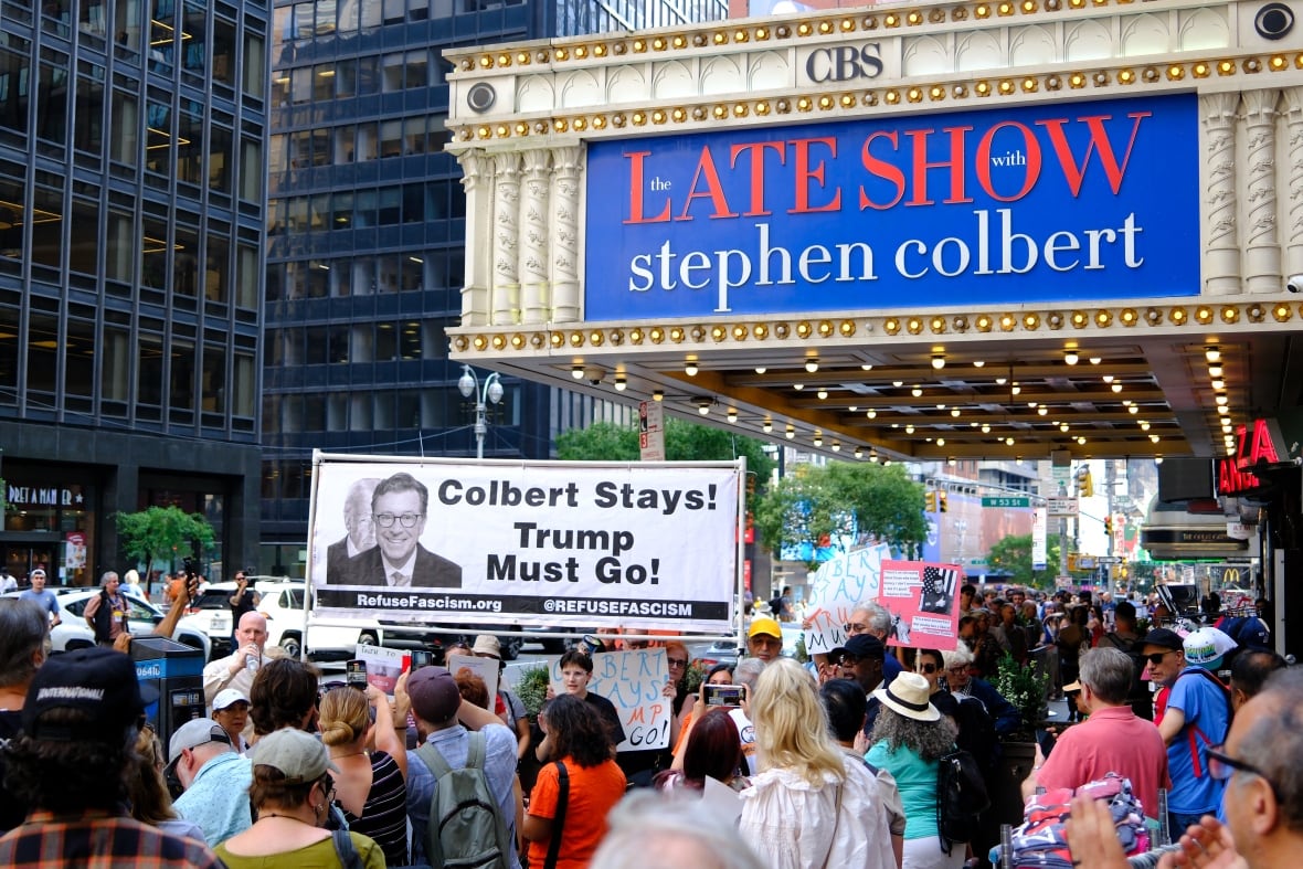 A crowd of protesters stand with a banner reading "Colbert Stays! Trump Must Go!" outside a theatres with a marquee displaying "CBS" and "The Late Show with Stephen Colbert."
