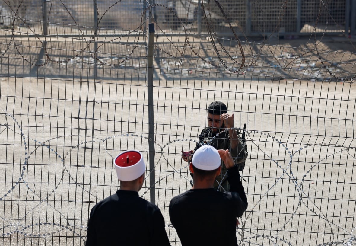 Two men speak to a soldier behind barbed wire.