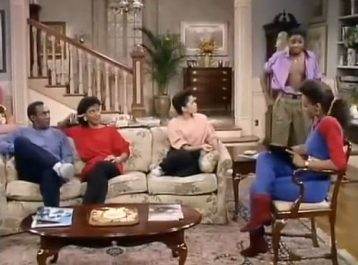 A still frame from an episode of The Cosby Show showing the inside of a home, with a large staircase in the background, with man, a woman and a young woman sitting on a couch, a young man standing to the right of the couch and to the left of a young woman sitting in a chair.