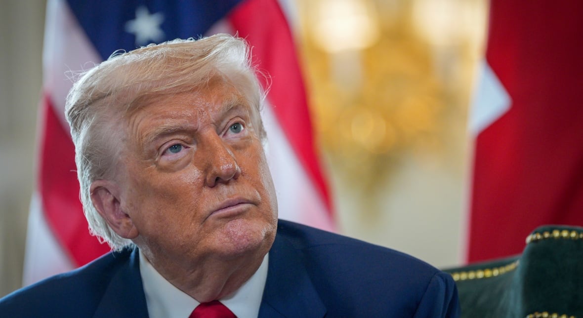 A man is shown from the shoulders up, looking upwards and into the distance. He is wearing a blue suit and there are flags behind him. 