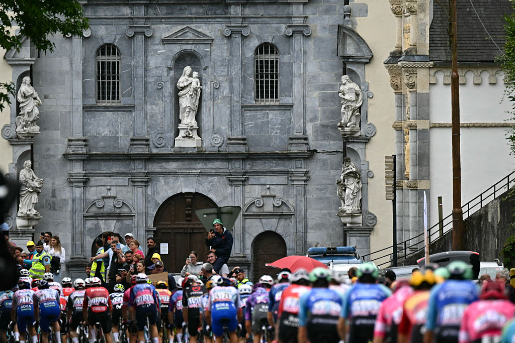The pack of riders (peloton) cycles past the Sanctuaire Notre-Dame de Betharram in Lestelle-Betharram during the 14th stage of the 112th edition of the Tour de France cycling race, 182.6 km between Pau and Luchon-Superbagneres, in the Pyrenees mountains of southwestern France, on July 19, 2025. (Photo by Loic VENANCE / AFP)