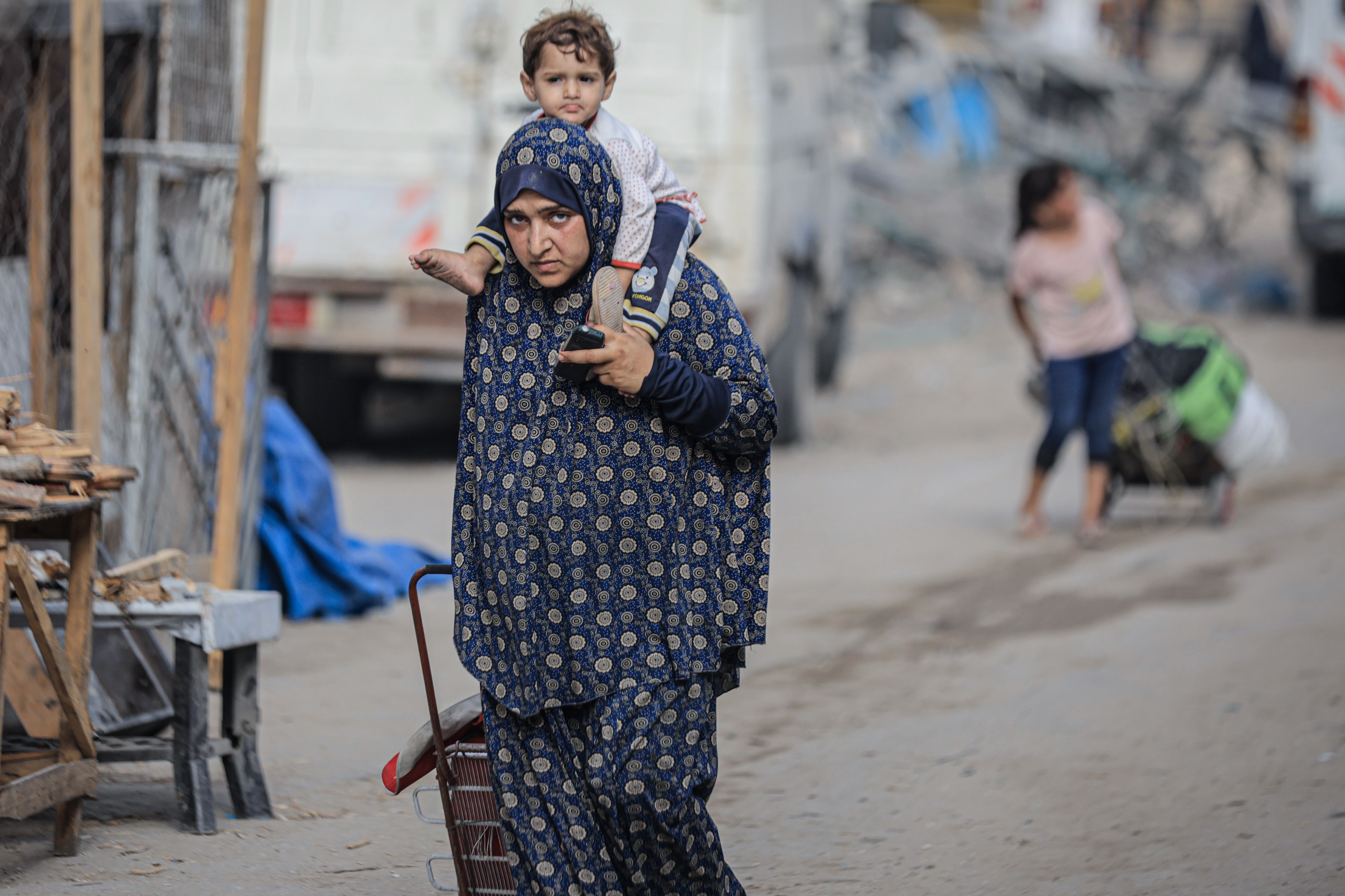 A Palestinian woman carries her child on her shoulders while pushing a cart, evacuating their home.