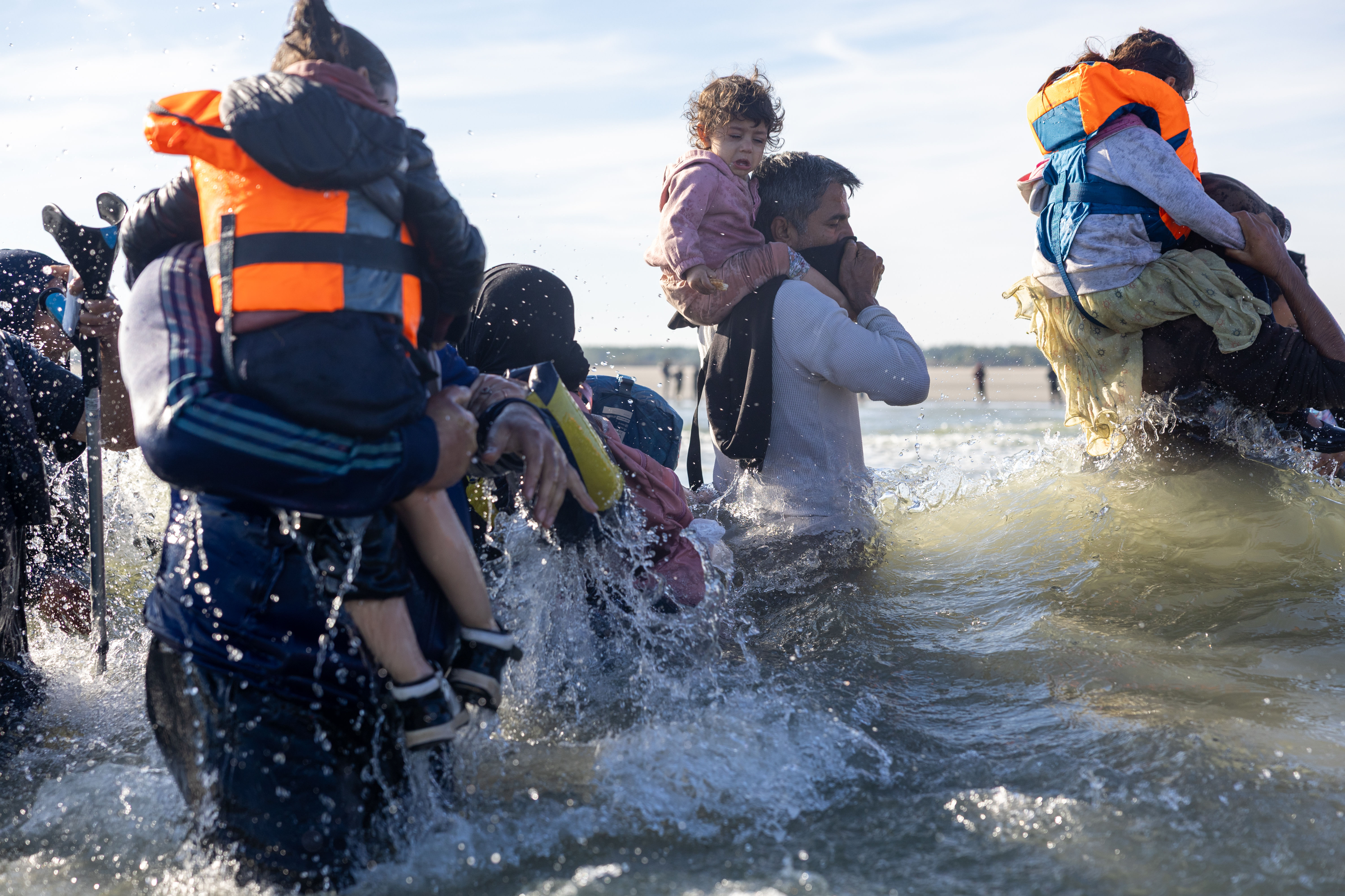 Migrant families wade through waves, children carried on shoulders, heading towards a small boat.