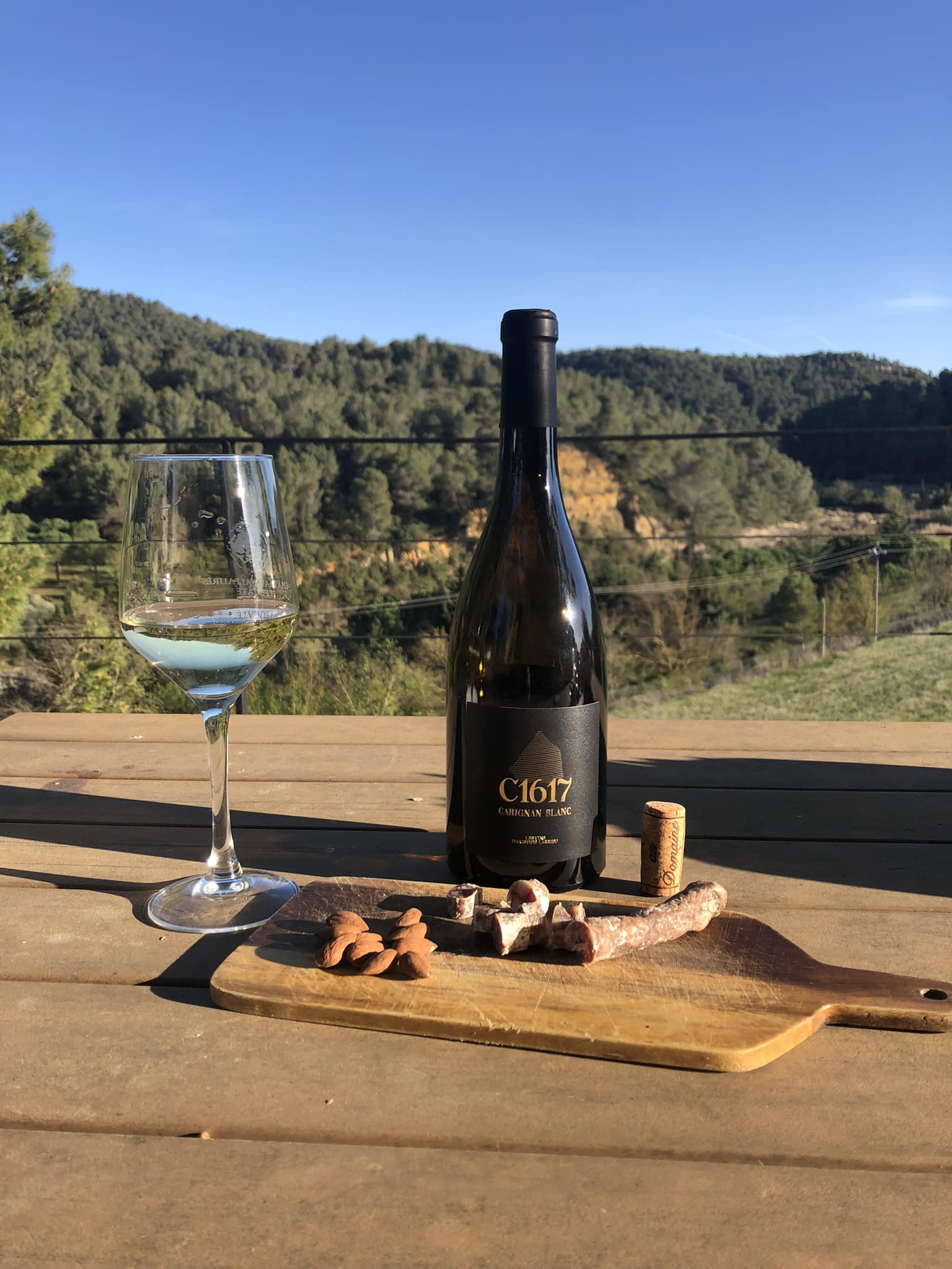 Bottle of C1617 Carignan Blanc wine, glass of white wine, almonds, and sausage on a wooden board outdoors.
