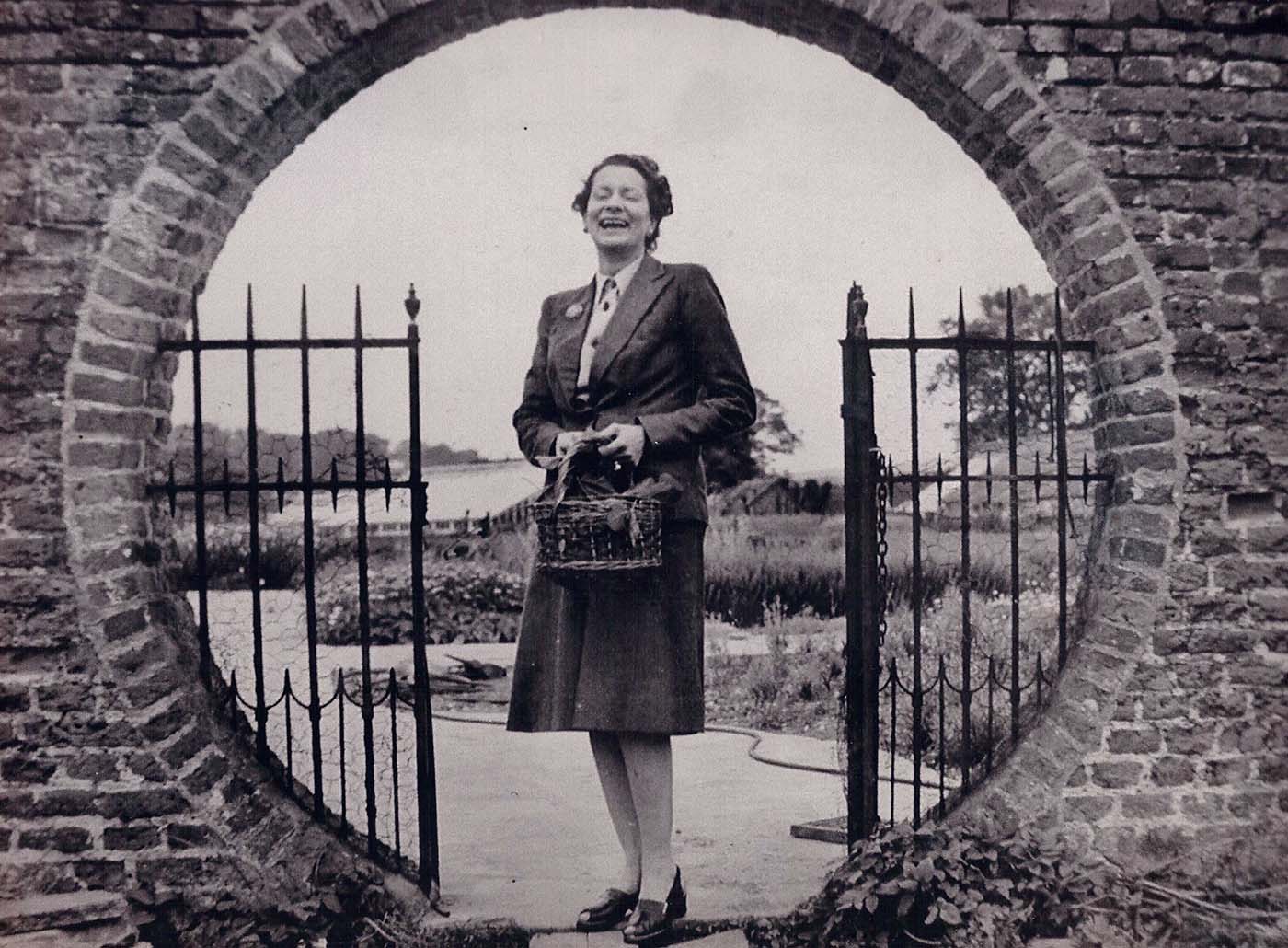 Black and white photo of Millicent Zoë, Lady Hart Dyke, smiling and holding a basket, standing in a circular brick archway.