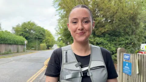 A woman wearing a black t-shirt and a grey jogging backpack. She is stood in front of a tree and a wooden fence, with a blue sign on it reading "EXIT".