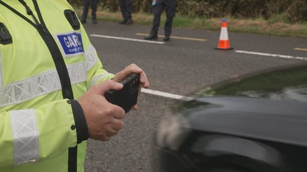 The hands of a garda holding a speed camera with a road, traffic cone and car in the background