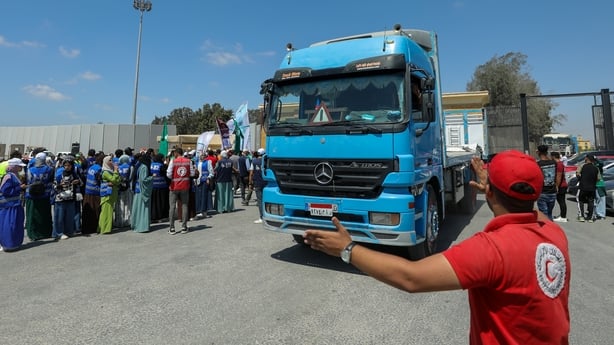 A man in a red tshirt and cap directs a bright blue aid truck down a road as dozens of people watch.