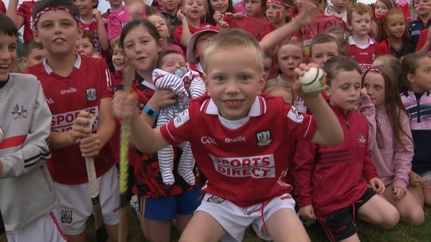 A young Cork fan holds up a sliotar and a hurl in front of a group of fellow fans