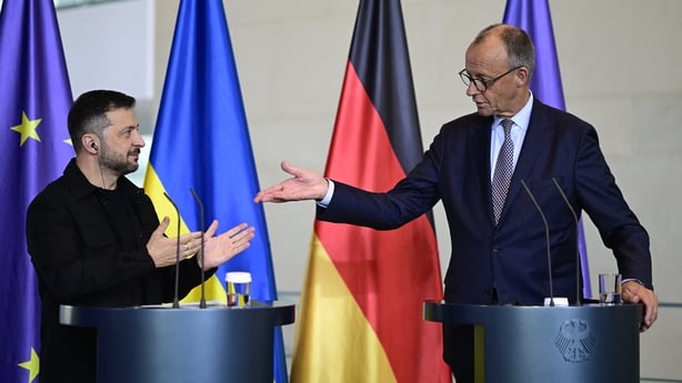 Two men standing at separate podiums gesture to each other with flags of germany and europe in the background