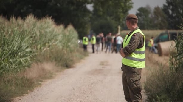 A man in army uniform and high vis vest looks at a group of people on a rural road