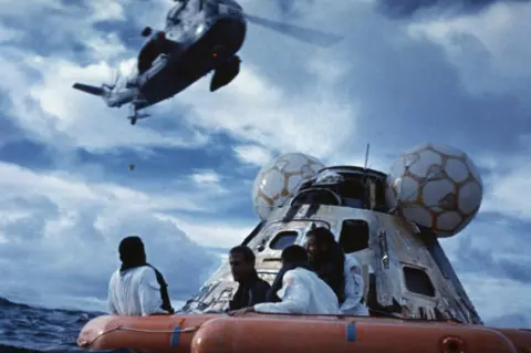 Getty Images Apollo 13 astronauts waiting for rescue in a raft.  The space capsule is shown behind them and a helicopter is hovering overhead.
