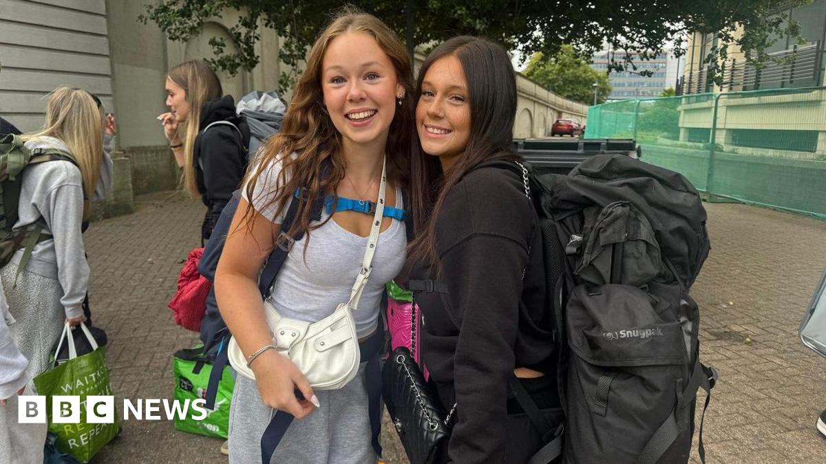 Two smiling young women with large backpacks pose for a photo while waiting in line, surrounded by other festival-goers with camping gear.