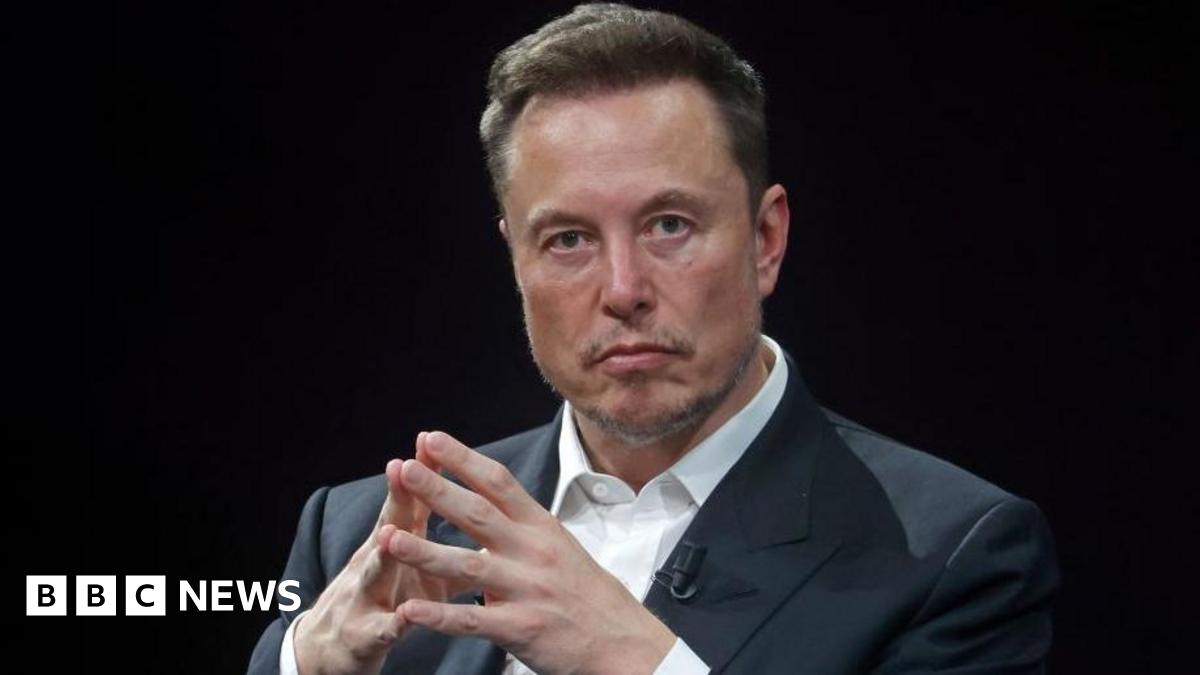 Elon Musk, who owned social media platform X, pictured with a stern expression, with his hands put together. He is dressed in a dark grey suit against a black backdrop.