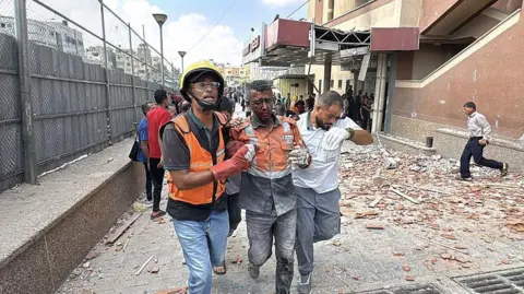 Getty Images An injured emergency worker is led away by two other emergency workers. There is rubble and wreckage behind them. He is badly hurt. 