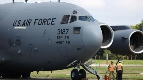 Getty Images A US Air Force plane sits in the runway. The nose of the grey aircraft and two engines on the left wing can be seen along with some ground crew in orange hi-viz jackets.