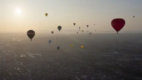 PA Numerous hot air balloons of all colours fly above Bristol at sunrise. The sky is hazy but clear. Hills and the River Severn can be seen in the distance.