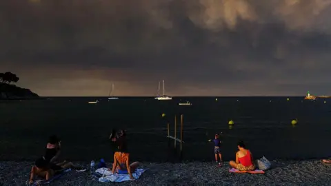 Getty Images The bottom of the image shows a number of people sat on a beach with the sea in front of them with a few boats and the sky filled with thick black smoke