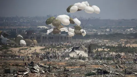 Getty Images Several parachutes with aid packages attached fall onto a barren landscape littered with ruined buildings. Hundreds of people move towards the aid packages. 