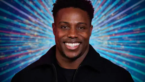 Harry Aikines-Aryeetey smiling in front of a multi-coloured glittery Strictly backdrop