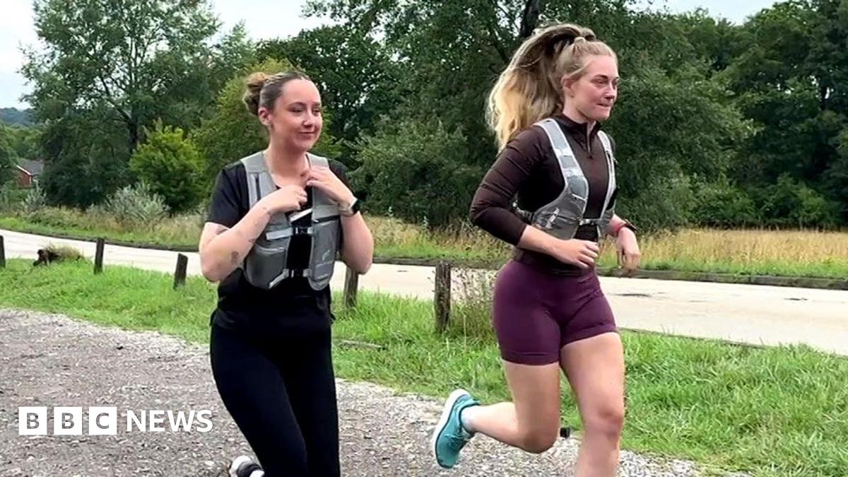 Image shows two females running. One is dressed in all black. She has a black short-sleeved top and black leggings on with a grey running vest over the top. She has black Nike trainers on. Her hair is in a bun. The second officer has turquoise trainers on. Burgundy shorts and a brown long-sleeved tight fitting top. She has a grey running vest over the top. Her hair is in a ponytail. They are in a carpark with four cars parked alongside them. Behind them are a number of trees, grass and a main road.