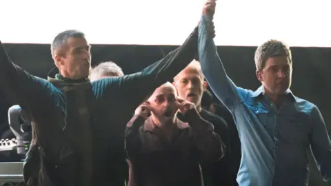 Getty Images Liam and Noel Gallagher with their arms raised aloft together while walking on-stage at their first comeback gig in Cardiff in July
