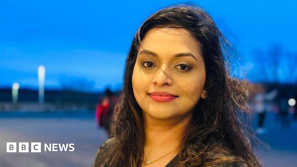 A woman with long dark hair is seen smiling at the camera. She has pink lipstick on and it is a headshot of her and a dark night.