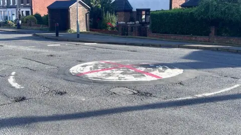 A red St George's cross painted on a white roundabout on a road with houses and shrubbery in the background.