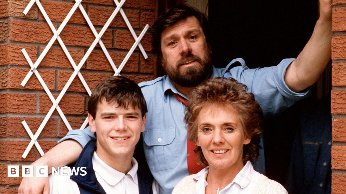 Bobby, portrayed by Ricky Tomlinson, and Sheila Grant, played by Sue Johnston are standing in front of their red-bricked home on Brookside Close with one of their children. They are all smiling at the camera in this posed photograph.