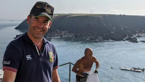 Mike Conner Mike Conner stands to the left of the photograph in front of the sea, he is wearing a blue polo shirt and dark cap and has short stubble. Behind him a man with no shirt on is walking up some steps carrying a bag. A small island can be seen in the background. 