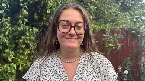 Gracie Moore A young woman wearing square, tortoiseshell glasses and a white and blue floral shirt smiles while standing in front of a tree laden with pears and a red wooden fence covered in vines.