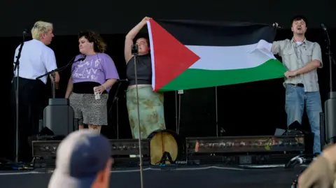 Getty Images Four members of The Mary Wallopers stood on the Victorious Festival stage holding a Palestinian flag and with mouths open appearing to chant