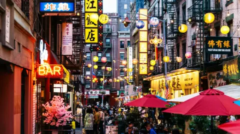 Getty Images A crowded street of restaurants and bars in New York City's Chinatown. Red, yellow and blue lanterns hang above the street